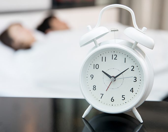 Alarm clock on a bedside table with two people sleeping in the background