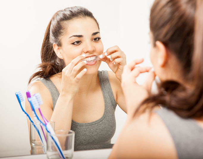 Woman in front of a mirror placing a whitening strip over her teeth